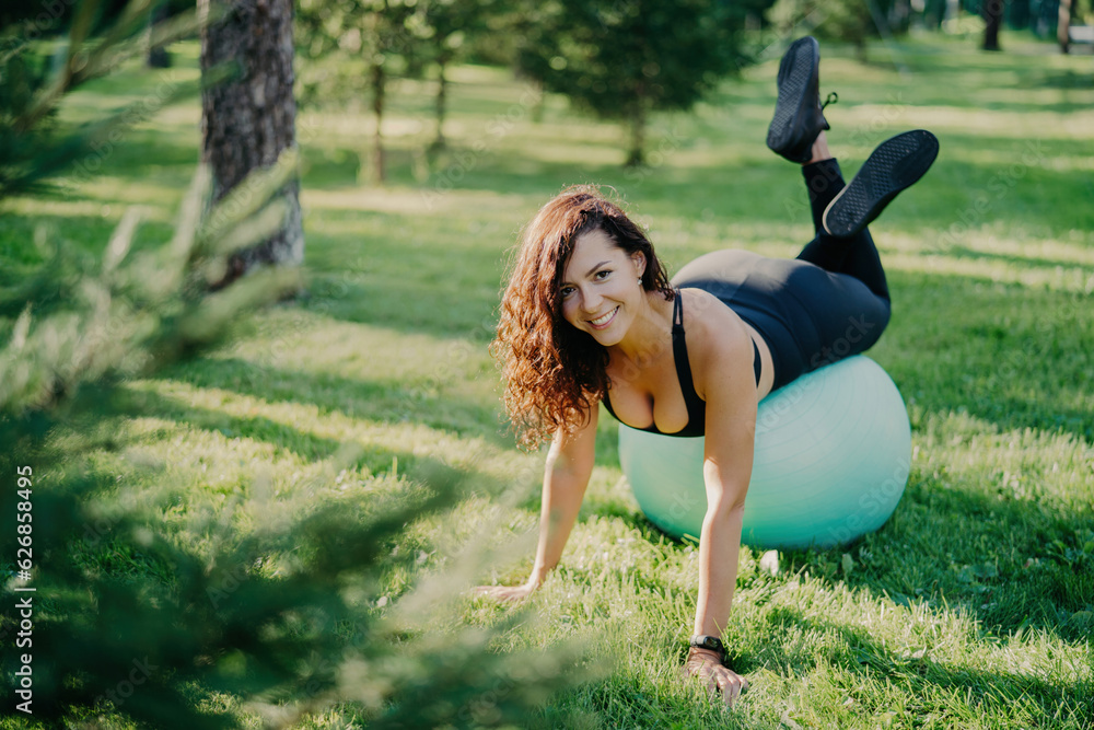 Fototapeta premium Smiling brunette exercises on green grass with fitness ball, aiming for a sporty body. Curly hair, sportswear, outdoor yoga training on a warm sunny day.