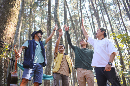 Smiling group of diverse young male friends standing at their forest campsite and raising hands with joy. Low angle view. Friendship, vacation and tourism concept photo