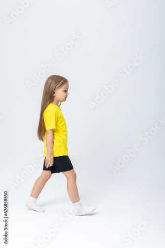 Calm and self-confident cute blonde kid girl in summer casual clothes walking over white background. Stylish comfortable everyday fashion for children concept