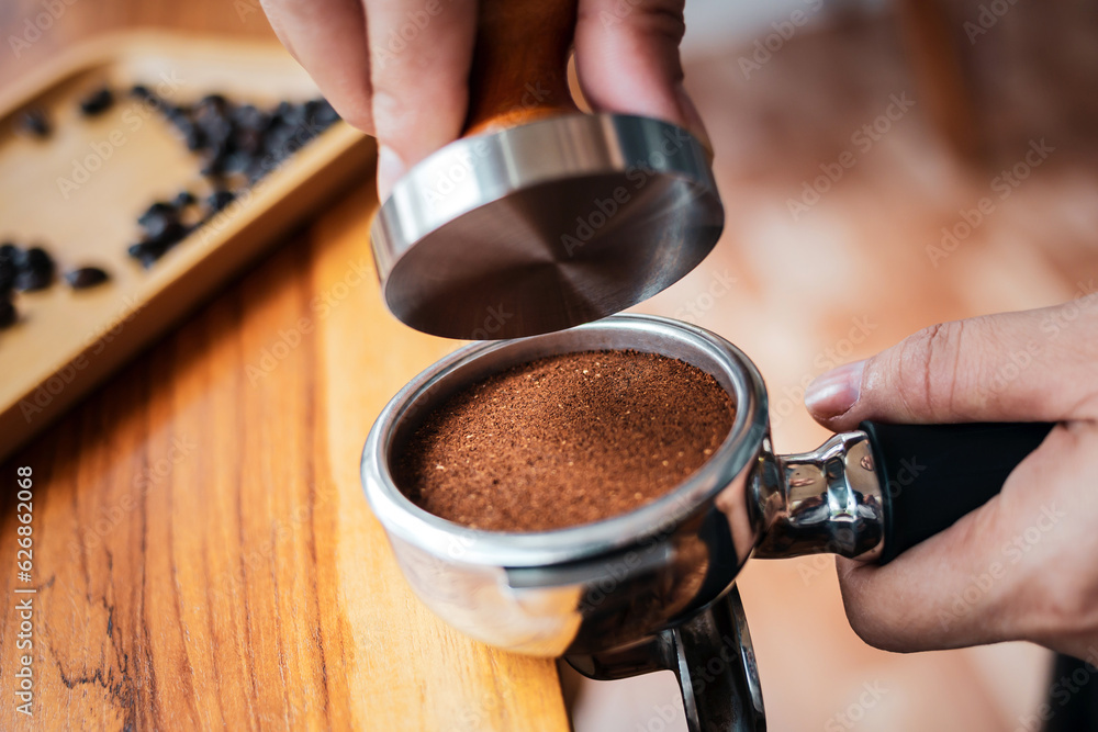 © NARONG - Close-up of hand Barista cafe making coffee with manual presses ground coffee using tamper on the wooden counter bar at the coffee shop