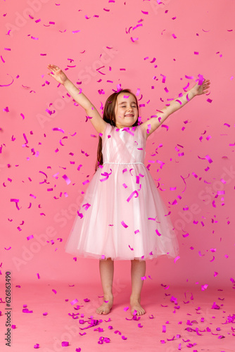 little girl in a pink dress catches confeti smiling happy on pink background, holiday concept. A child is celebrating a birthday on a pink background