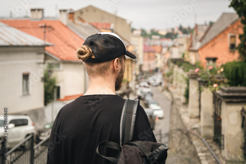 A man tourist in a cap, with a bun of hair and with a backpack explores the city looking at the panoramic view of the city, view from the back, copy space.