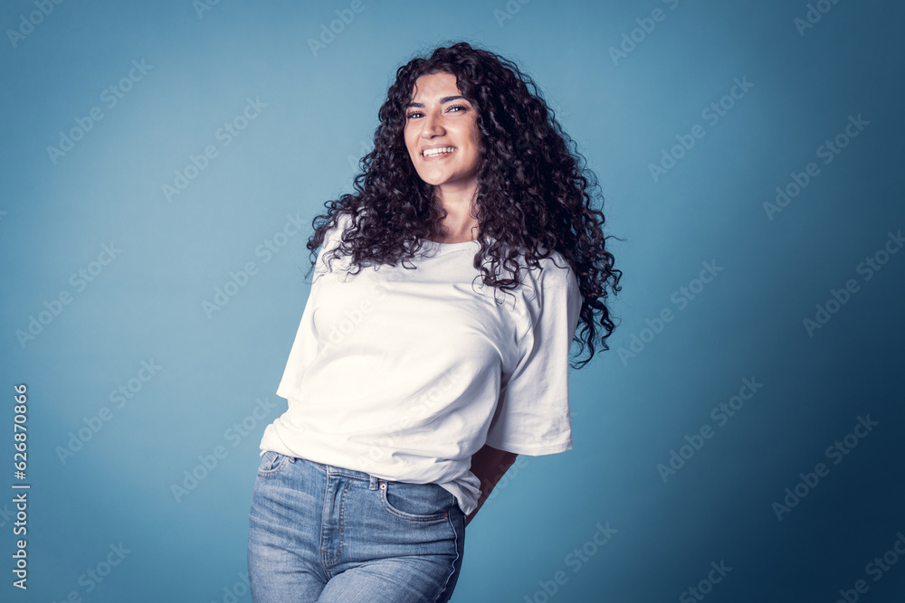 Shot of happy confident curly woman with toothy smile, wears casual basic solid white t-shirt, expresses good emotions, enjoys nice day, isolated over blue background. Expressions Shot of happy confident curly woman with toothy smile, wears casual basic solid white t-shirt, expresses good emotions, enjoys nice day, isolated over blue background. Expressions