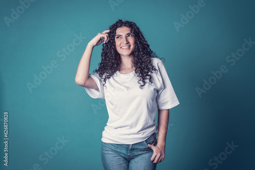 Shot of happy confident curly woman with toothy smile,wears casual basic solid white t-shirt, expresses good emotions, enjoys nice day, thinking isolated over blue background. Expressions © Diego Zarulli