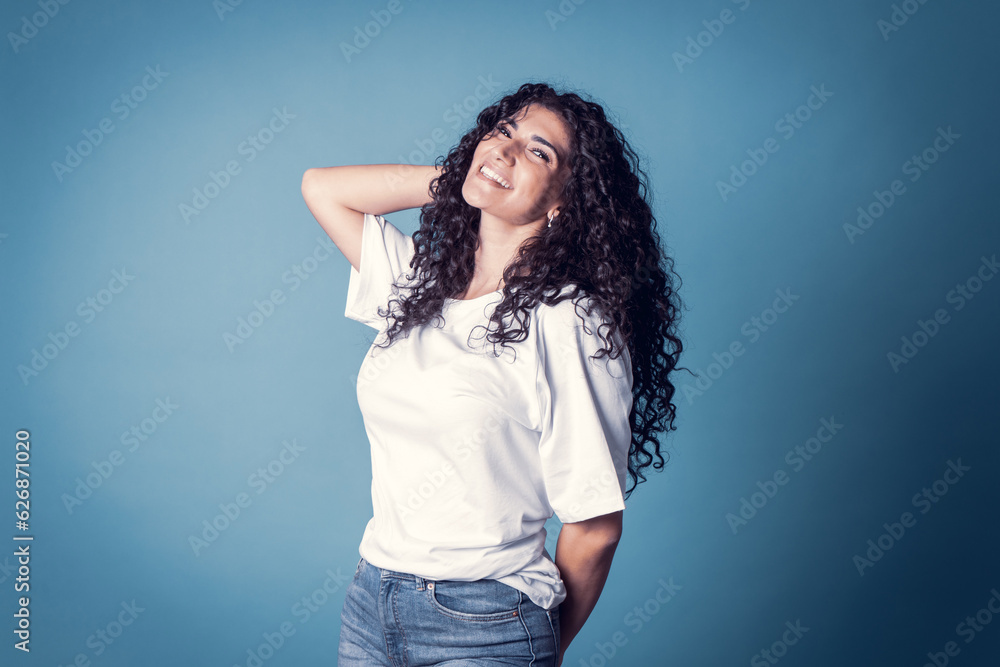 Shot of happy confident curly woman with toothy smile, wears casual basic solid white t-shirt, expresses good emotions, enjoys nice day, isolated over blue background. Expressions Shot of happy confident curly woman with toothy smile, wears casual basic solid white t-shirt, expresses good emotions, enjoys nice day, isolated over blue background. Expressions