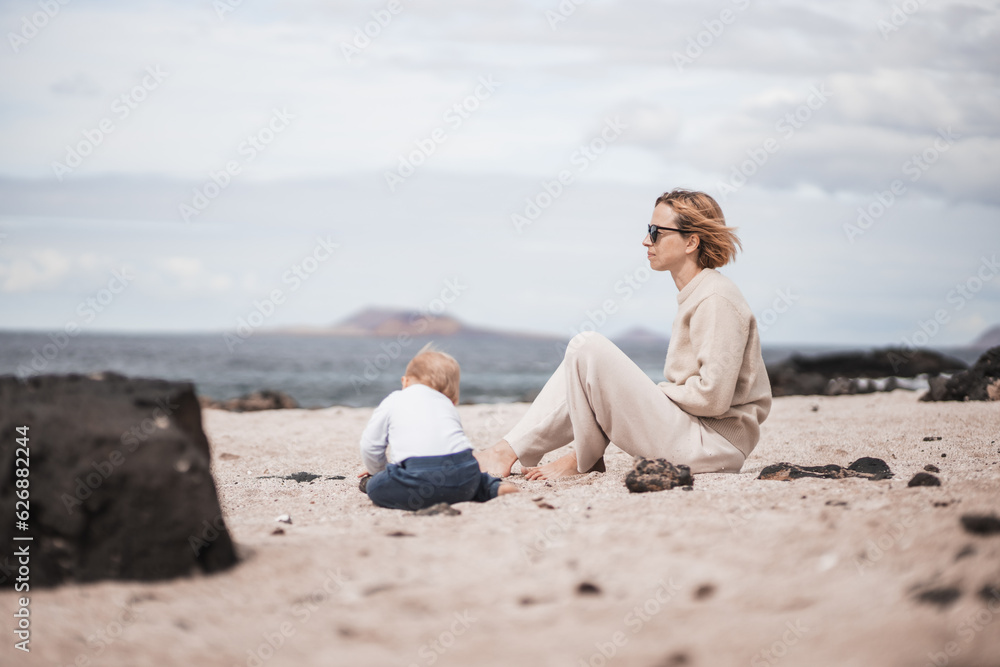 Mother enjoying winter beach vacations playing with his infant baby boy son on wild volcanic sandy beach on Lanzarote island, Canary Islands, Spain. Family travel and vacations concept.