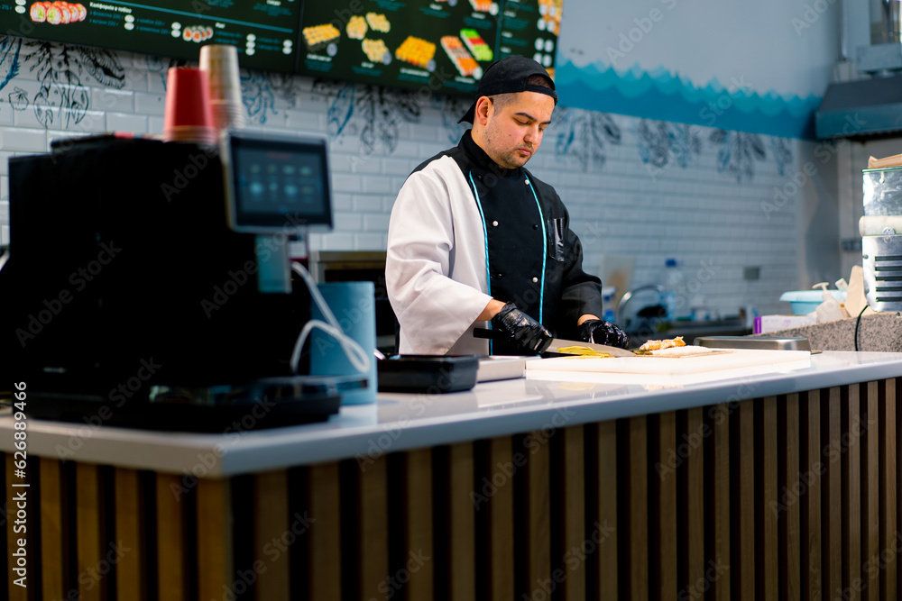 Fototapeta premium Sushi chef in the process of preparing a philadelphia roll with seared salmon cream cheese and cucumber on a white kitchen board