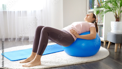 Young woman in leggings stretching back muscles while rolling on blue fitness ball