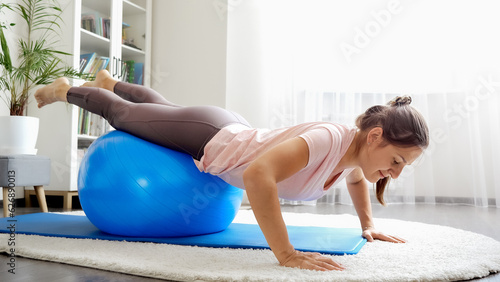 Smiling young woman doing exercises and balancing on fitball at living room. Concept of healthcare, sports and yoga at home.