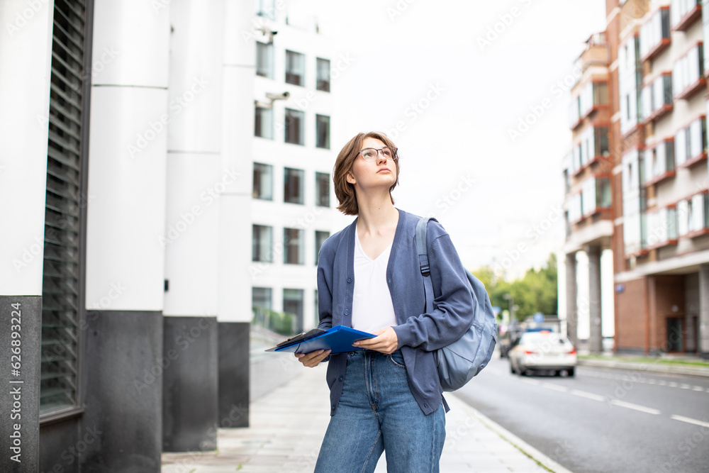 Young blonde woman talking on a mobile phone, smiling, holding folder and looking to camera on the background of an office building. Corporate employee. IT-specialist