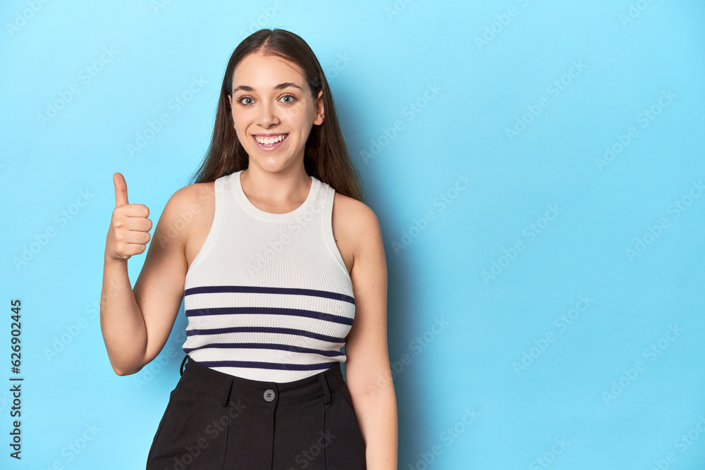 Woman in a striped top posing on a blue studio backdrop smiling and raising thumb up Woman in a striped top posing on a blue studio backdrop smiling and raising thumb up