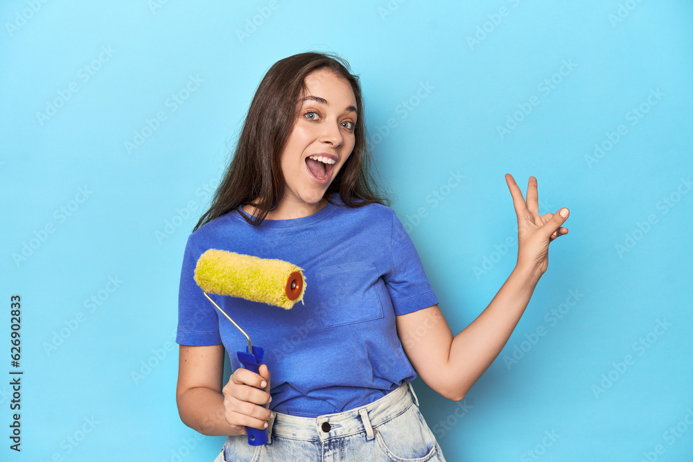 Young woman with yellow paint roller on a blue background joyful and carefree showing a peace symbol with fingers. Young woman with yellow paint roller on a blue background joyful and carefree showing a peace symbol with fingers.