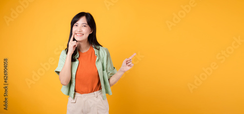 dental health with captivating young Asian woman 30s, donning orange shirt and green jumper points to her teeth, isolated on vibrant yellow background, emphasizing the importance of dental healthcare.