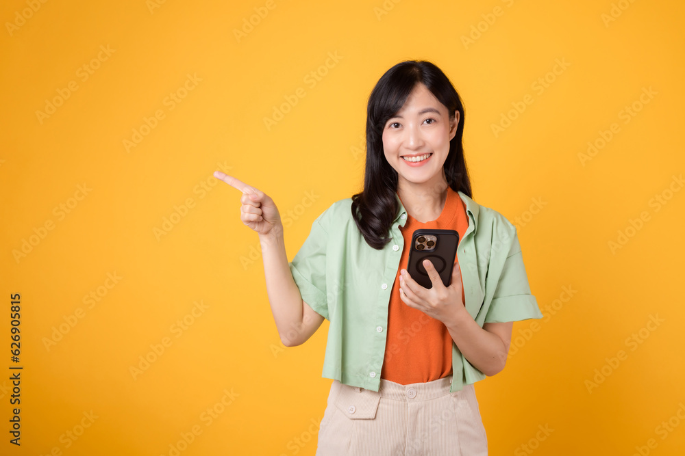 digital era come alive with young Asian woman 30s wearing orange shirt presents a smartphone with a finger hand gesture. The isolated yellow studio background adds charm to the app smartphone concept.