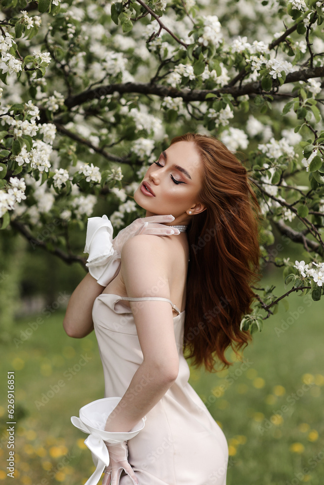 Red-haired girl in a lite dress in blooming apple trees. The girl poses against the background of flowers in the spring park. Red-haired girl in a lite dress in blooming apple trees. The girl poses against the background of flowers in the spring park.