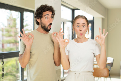 young adult couple feeling shocked  amazed and surprised  showing approval making okay sign with both hands