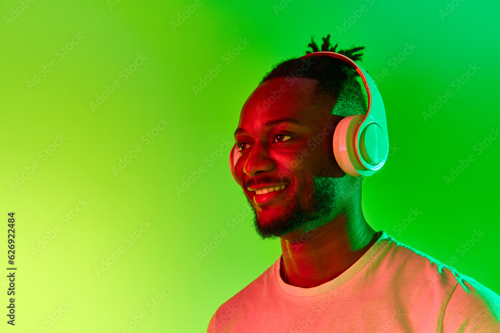 Image of young african man looking away in headphones and smiling over gradient colorful background in neon. Image of young african man looking away in headphones and smiling over gradient colorful background in neon.