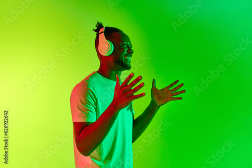 Annoyed young man with dreadlocks looking away in headphones with raising hand up over gradient colorful background in neon. photo