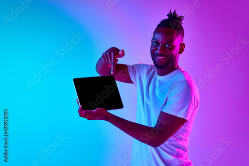 Young african man with afro hairdo holding padlet pointing at gadget screen and looking at camera over gradient blue-pink background in neon. photo