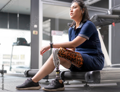 Woman with prosthetic leg sits isolated in gym. Asian female with foot prosthesis physical exercise in fitness. Artificial limb equipment help accident survivor or body injury people to mobility.