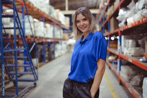 Confident female warehouse worker standing in logistic storage looking at camera smiling. Portrait of young woman employee working in distribution warehouse business. Industrial staff in storehouse.
