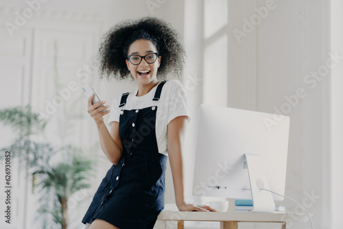 Joyful female financier, mobile in hand, salary received on time, by wooden table, working on computer, wearing white t-shirt and sarafan, happily texting. photo