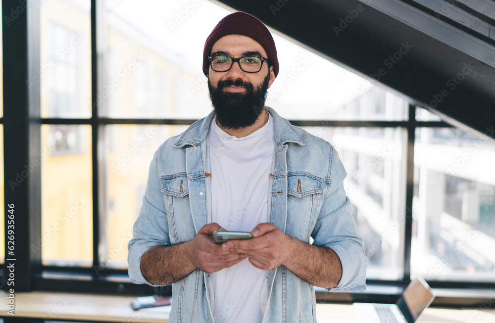 Smiling hipster browsing cellphone standing against big window Smiling hipster browsing cellphone standing against big window