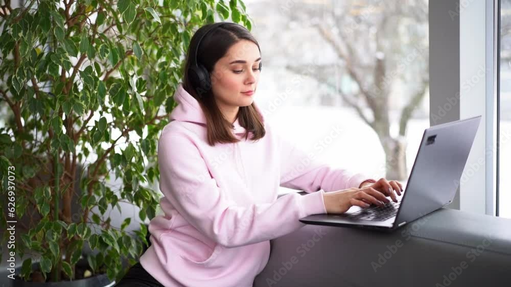 A girl in headphones sits near the window, works and communicates online by the laptop in a cafe A girl in headphones sits near the window, works and communicates online by the laptop in a cafe