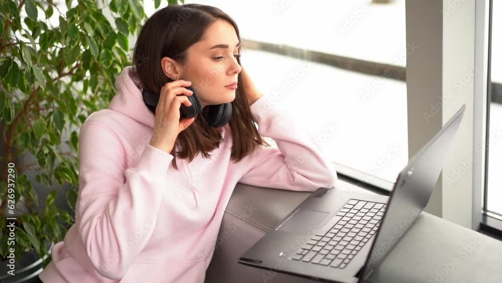 A girl in sweatshirt and headphones sits by the window, she is listening the music, works and communicates by the laptop the cafe A girl in sweatshirt and headphones sits by the window, she is listening the music, works and communicates by the laptop the cafe