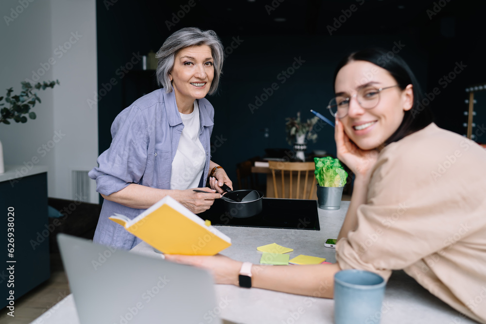 Cheerful mature woman standing with coffee pan at kitchen while daughter joining with recipe
