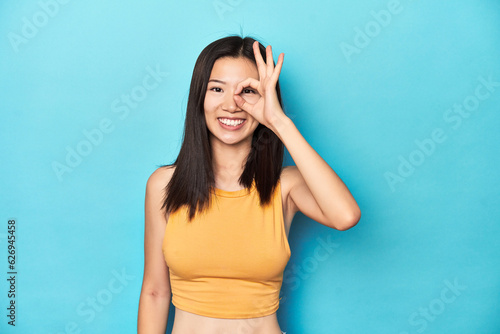Asian woman in summer yellow top, studio setup, excited keeping ok gesture on eye.