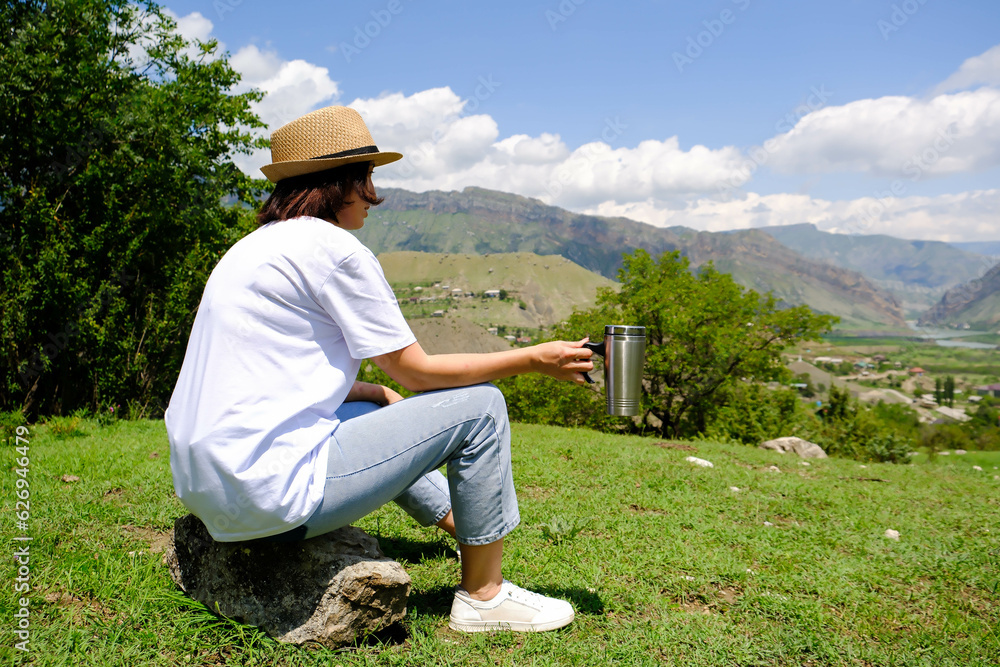 A tourist girl sits on a stone and drinks tea from a thermos on top of the mountains.