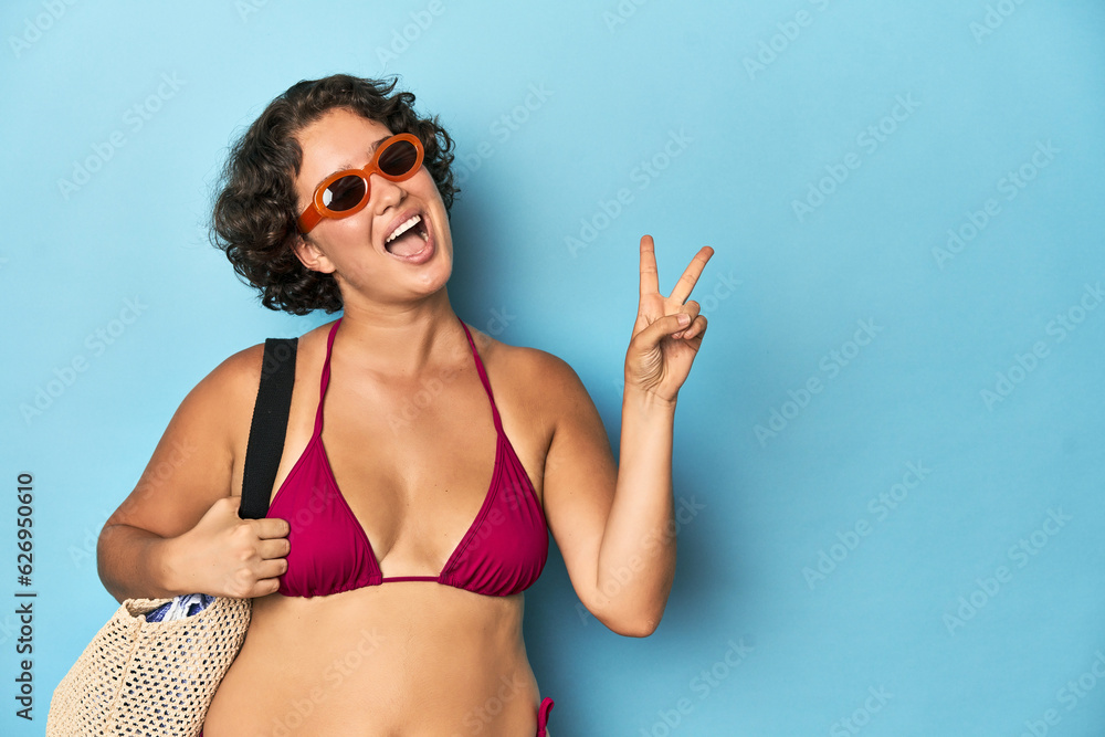 Young woman in bikini with beach bag joyful and carefree showing a peace symbol with fingers.