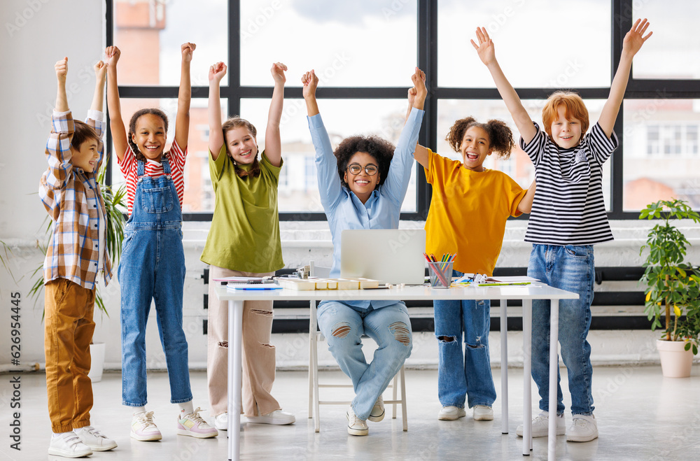 Fototapeta premium Teacher and cheerful schoolkids looking at laptop screen and celebrate successful completion of collective school work during online lesson