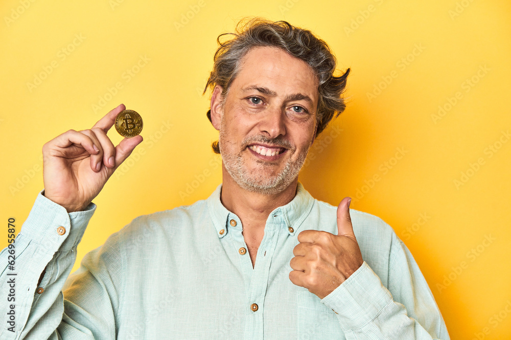 Man holding a physical bitcoin, yellow backdrop smiling and raising thumb up Man holding a physical bitcoin, yellow backdrop smiling and raising thumb up
