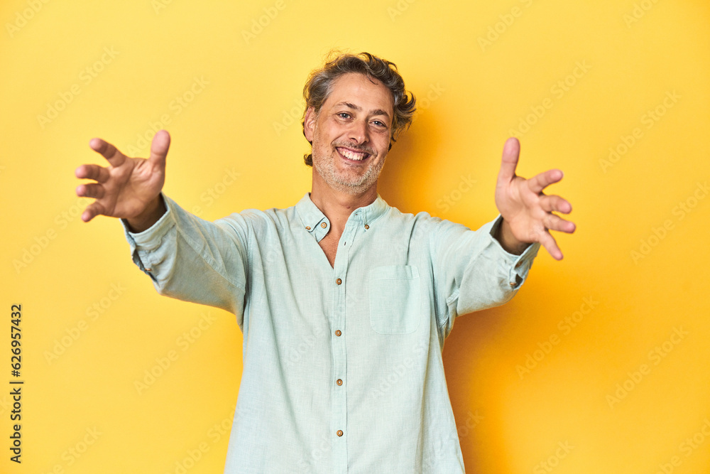 Middle-aged man posing on a yellow backdrop feels confident giving a hug to the camera.