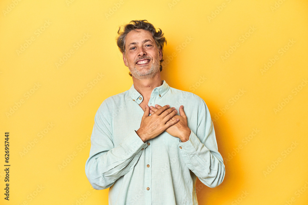 Middle-aged man posing on a yellow backdrop laughing keeping hands on heart, concept of happiness.