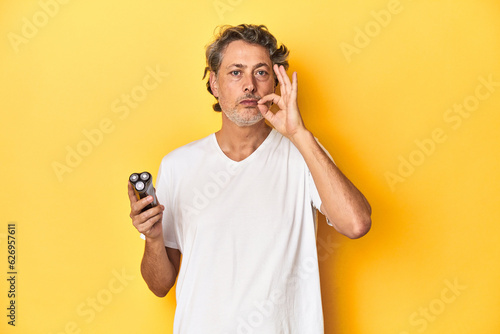 Man holding a razor, yellow studio background with fingers on lips keeping a secret. © Asier