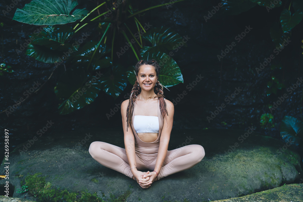 Obraz premium Portrait of cheerful young woman practicing hatha at beautiful nature environment with leaves on background, happy relaxed fit girl performing yoga routine while sitting in Lotus pose outdoors