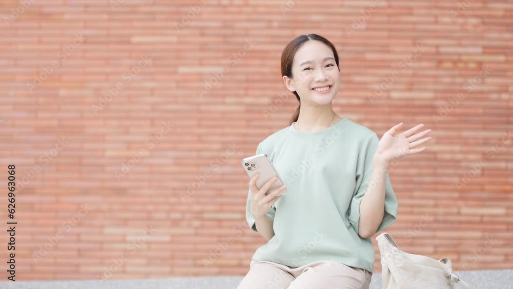 Asian woman greets camera holding smartphone while sitting in university Asian woman greets camera holding smartphone while sitting in university