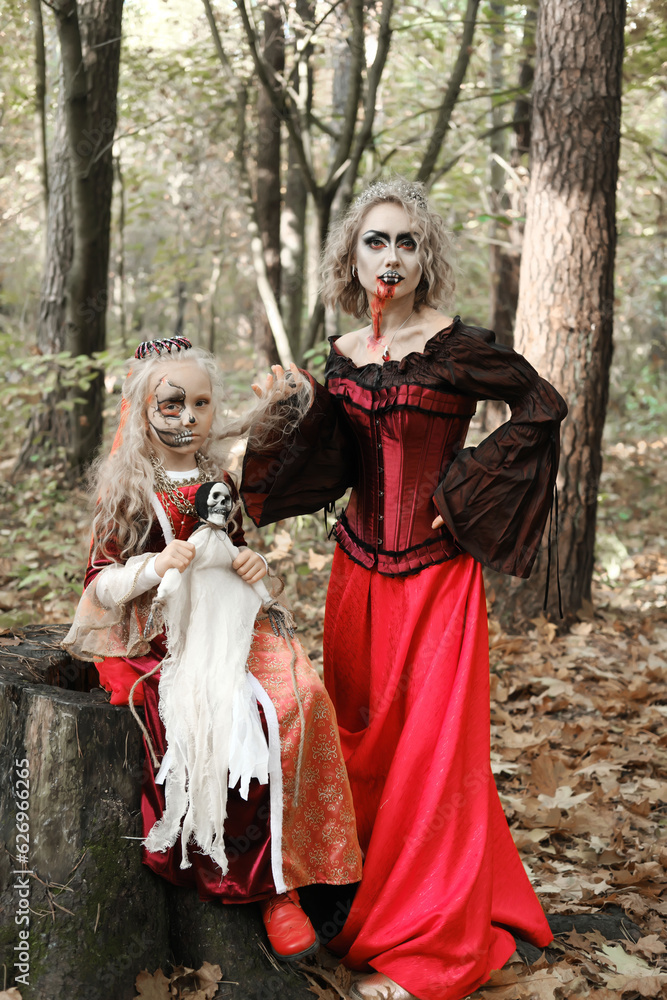 A woman in the form of a vampire or a sorceress and a girl in the form of a dead princess with a skeleton doll in their hands pose in the forest. Models are looking at camera A woman in the form of a vampire or a sorceress and a girl in the form of a dead princess with a skeleton doll in their hands pose in the forest. Models are looking at camera