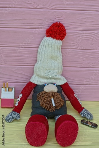 toy beautiful homemade original funny in a white hat with a red balabon brown beard and red shoes a childish gnome smokes a cigarette with a yellow filter and lies on a yellow table  photo