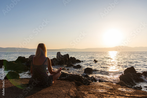 Beauty young woman in dress sitting on rock in yoga pose near sea at sunset. Fitness, meditation background, Croatia