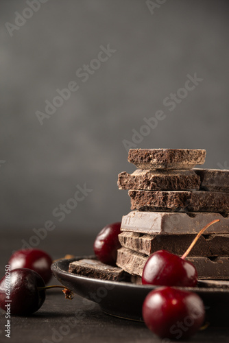 View of stack of chocolate bars in plate on wooden table with cherries, selective focus, gray background, vertical, with copy space