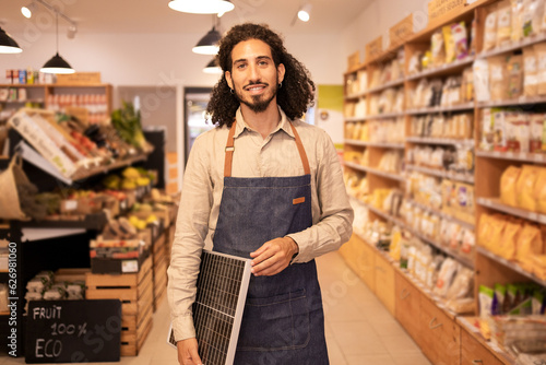 Cheerful ethnic seller holding portable solar panel in store photo