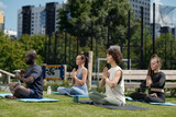 Side view of group of intercultural athletes practicing yoga outdoor while sitting on mats with their legs crossed and palms put together by chest