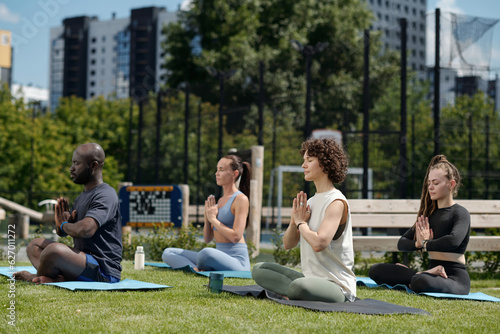 Side view of group of intercultural athletes practicing yoga outdoor while sitting on mats with their legs crossed and palms put together by chest