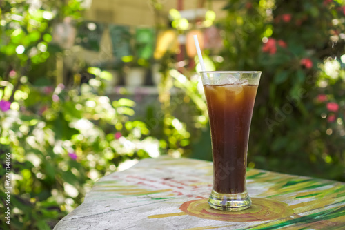 Iced Americano or Long Black Coffee in a long glass with a straw on a wooden table against a bright bokeh garden background. Concept for working, task, job, business plan, and outdoor cafe.