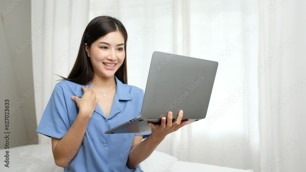 A pleasant young woman smiling and waving her hand during a video call on a modern laptop in bed. Happy lady having online communication during evening time at a hotel room. A pleasant young woman smiling and waving her hand during a video call on a modern laptop in bed. Happy lady having online communication during evening time at a hotel room.