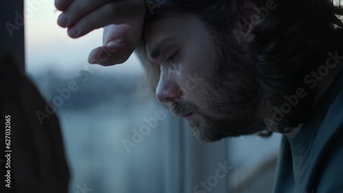 Depressed man crying, thinking about problems near window. Close up of pensive, sad depressed unhappy beard man with long hair. Mental health, man in tears, man cry depression concept. Authentic